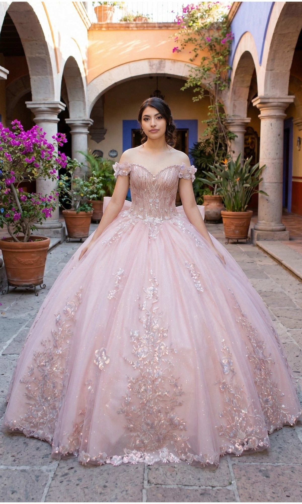 A young woman stands in a courtyard wearing the Quinceanera Dress 8370J by Cinderella Couture, a blush pink ball gown featuring an off-shoulder neckline, floral appliques, and a full skirt.